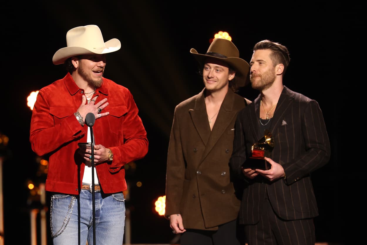 A man in a red jacket and cowboy hat holds a phone in one hand while speaking into a microphone, on stage during the Grammy awards. 