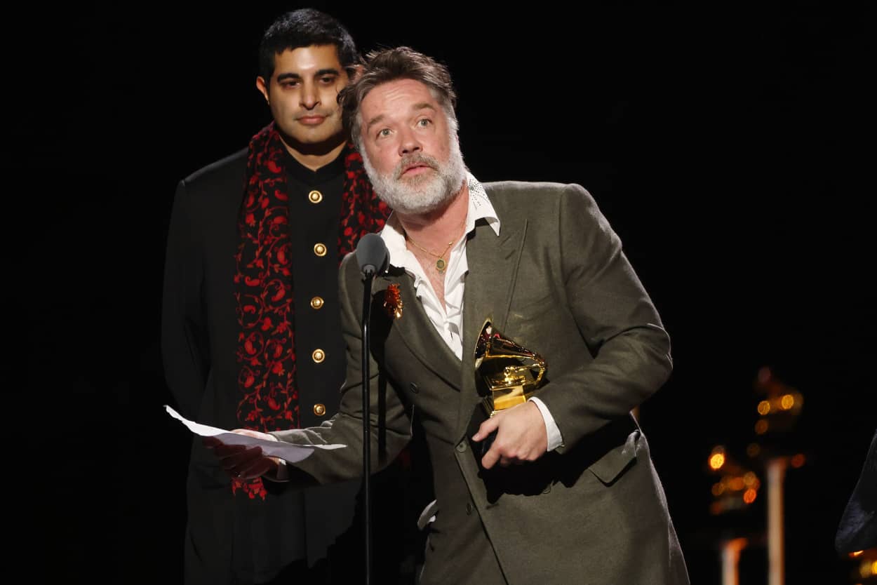 Rufus Wainwright holds a golden Grammy award in his left hand while leaning into a microphone, on stage at the Grammy awards. 