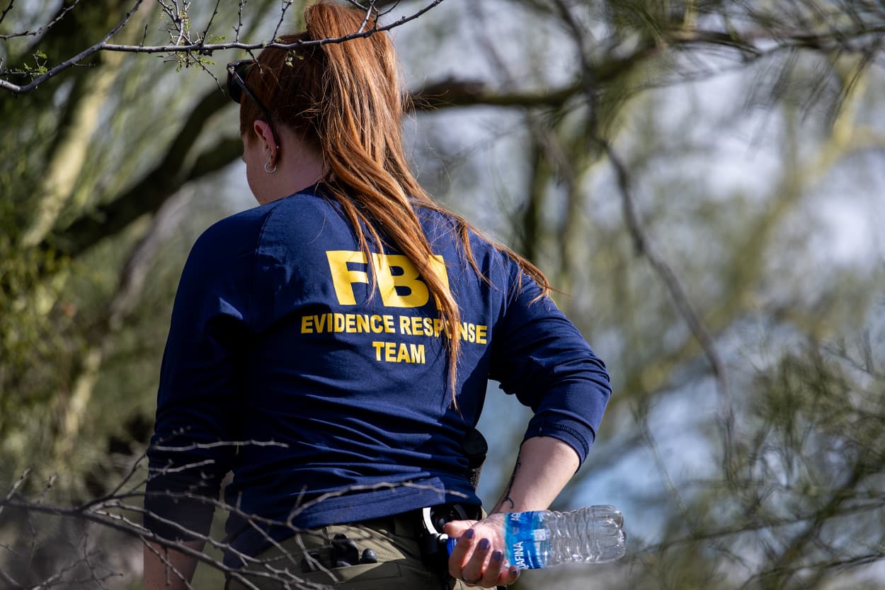 A woman with a long ponytail and a blue longsleeved shirt that says FBI EVIDENCE RESPONSE TEAM walks among trees.