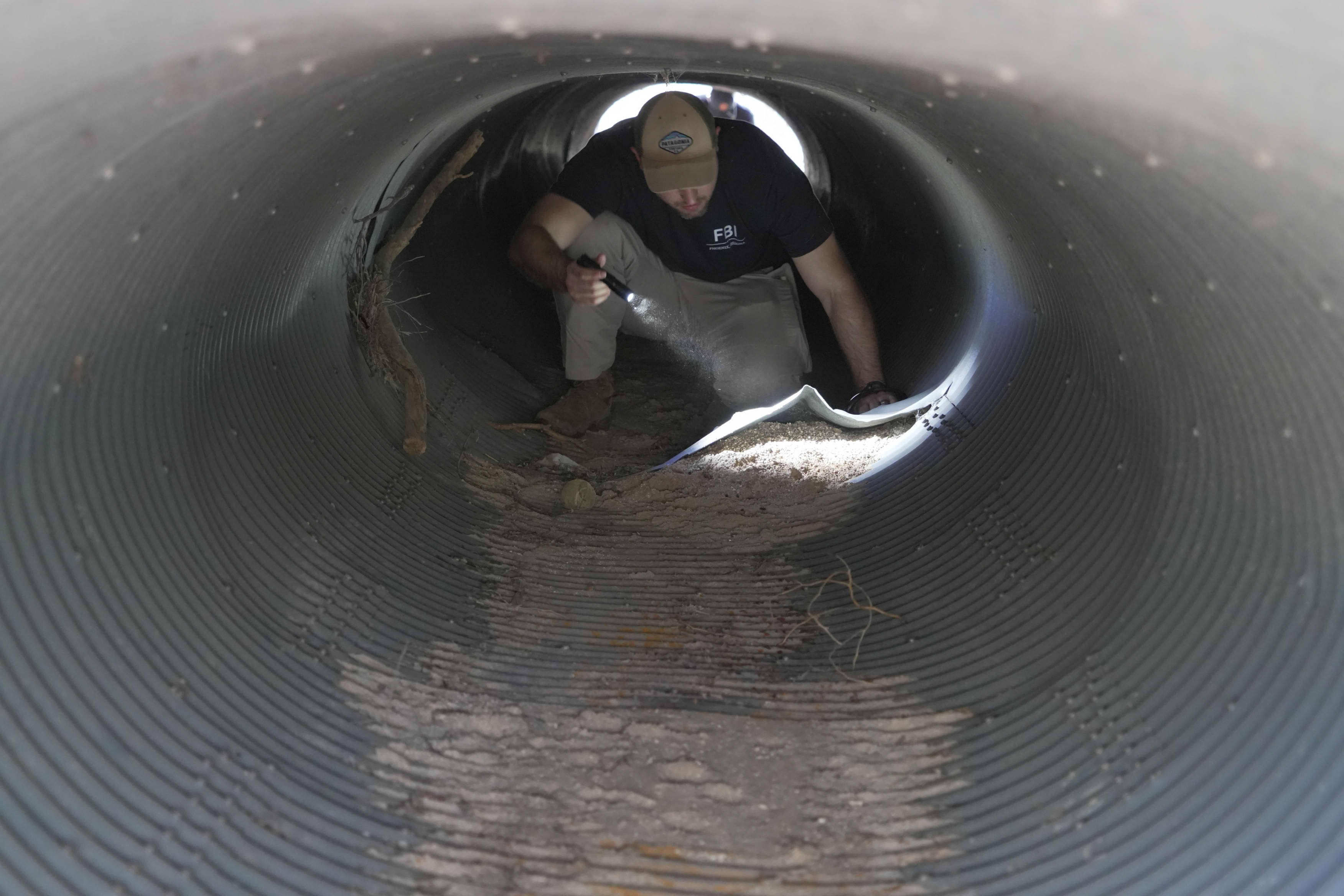 A man wearing an FBI t-shirt and holding a flashlight crawls on his hands and knees in a metal culvert.