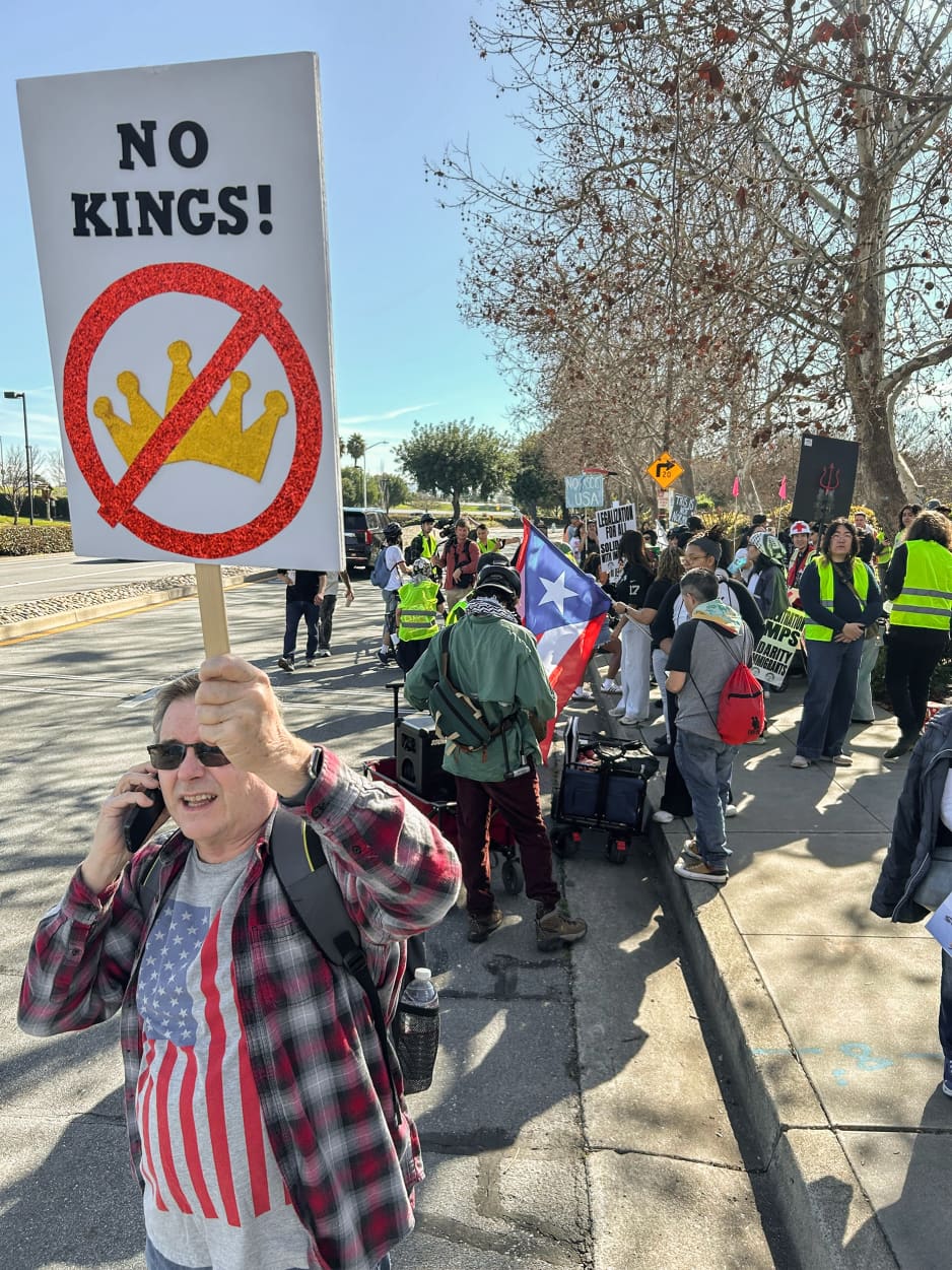  A man wearing sunglasses and holding a NO KINGS sign in the air speaks on his phone during a roadside demonstration. 