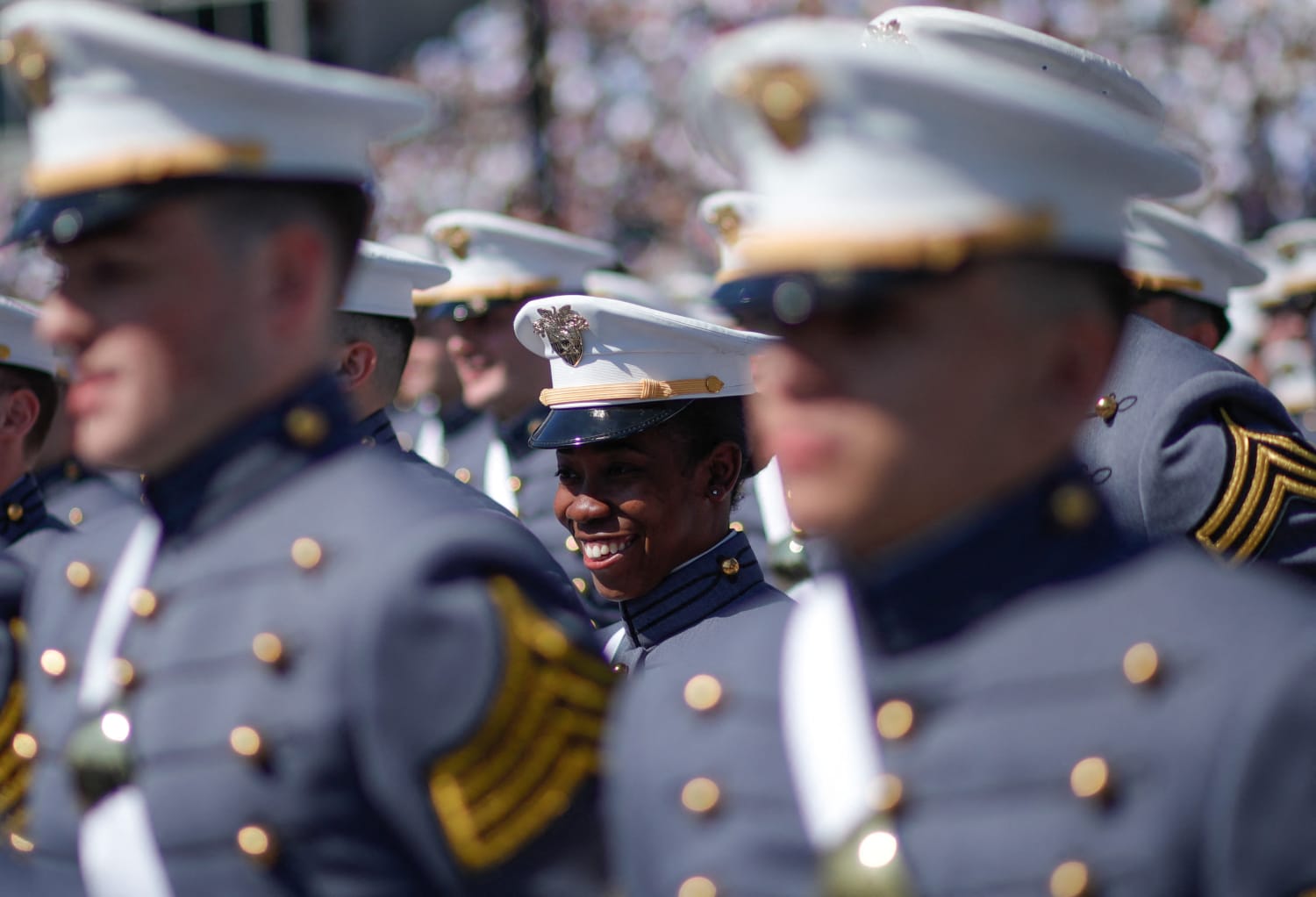 A sea of military personnel adorned in their celebratory garb with their faces faded out besides one smiling, young Black woman in the center of the frame.