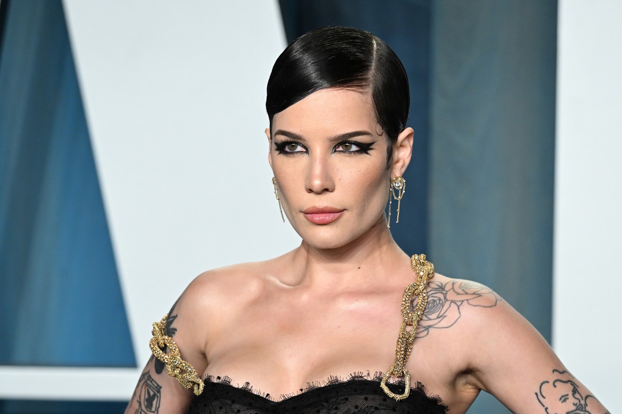A woman with brown hair is wearing an elegant black dress at an official event. On her shoulder and arm are several tattoos.