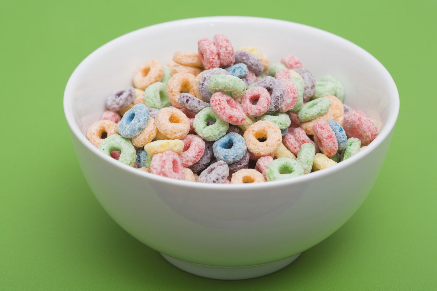 The image showcases a vibrant and appealing bowl of colorful cereal against a solid, bright green background. The cereal appears to be Fruit Loops, characterized by its small, circular, ring-shaped pieces in a variety of hues – red, orange, yellow, green, blue, and purple. The bowl itself is a classic, rounded white ceramic design, sitting slightly elevated on a small footed base. It’s generously filled with the cereal, creating a mound that nearly reaches the rim. The texture of the cereal looks slightly frosted or sugared, catching the light and adding to its visual appeal. The green backdrop provides a strong contrast to the colorful cereal, making the colors pop and drawing the viewer's eye directly to the bowl. The lighting is even and bright, highlighting the details of both the cereal and the bowl. There are no other objects or elements present in the image, keeping the focus solely on the cereal and its presentation. The overall composition is simple yet effective, evoking a sense of childhood nostalgia and a cheerful breakfast experience. The image is well-focused and clear, allowing for a detailed observation of the cereal’s shape, color, and texture.