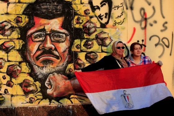 Image: Women hold the Egyptian national flag as they stand in front of graffiti depicting President Mohammed Morsi outside the presidential palace in Cairo.