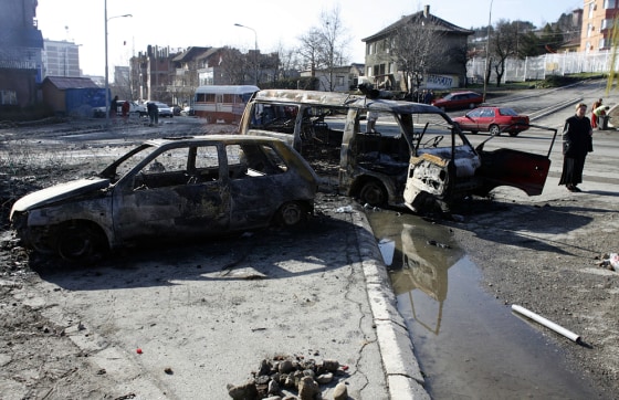 Image: A woman walks by burnt vehicles in the ethnically divided city of Mitrovica