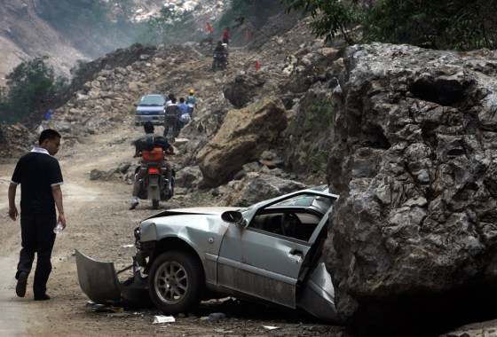 Image: A man looks at a car being half flattened by a huge rock as motorists ride through a damaged road