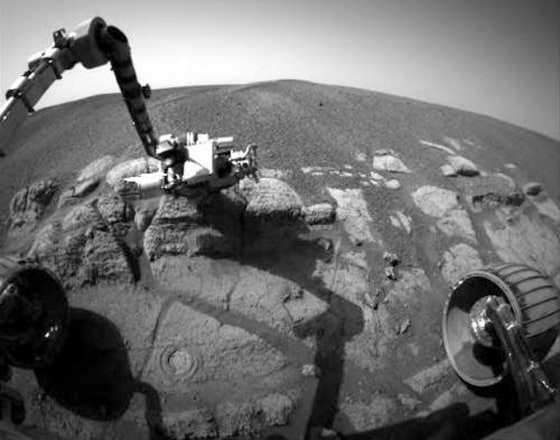Opportunity's hazard avoidance camera provides a view of its robotic arm placed on a bedrock outcropping for an analysis of the rock. A hole previously drilled in the outcrop is visible toward the bottom of the image, and one of the rover's front wheels is in the lower right corner.