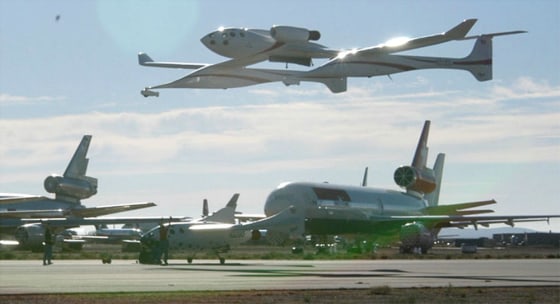 Scaled Composites' White Knight carrier aircraft makes a low pass over the SpaceShipOne rocket plane, surrounded by other planes parked at the Mojave Airport in California.