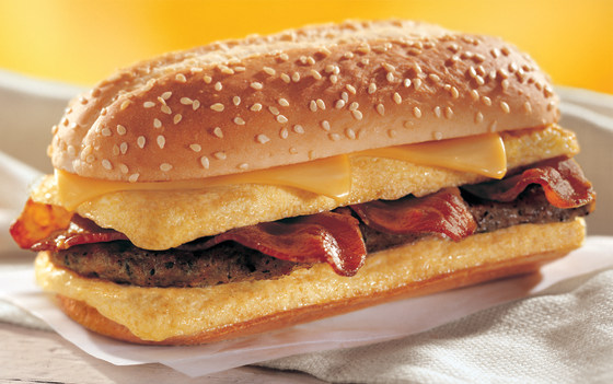 The "Enormous Omelet Sandwich" contains two slices of melted, American cheese, two fluffy eggs stuffed with three crispy strips of bacon, and a sausage patty, piled high on a toasted bun.