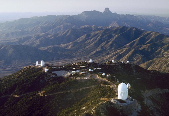 An aerial view shows Kitt Peak's telescopes in the foreground and Baboquivari Peak in the background, as a knob rising dramatically from the surrounding ridge of mountains.