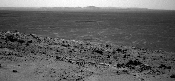 A picture from the Spirit rover's navigational camera shows terrain leading up to the summit of Husband Hill in the foreground, with what appears to be a crater on the plain below and distant hills on the horizon.