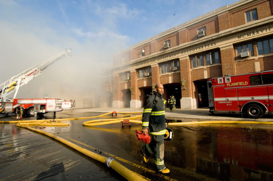 A firefighter walks in front of a burning mattress supply building in Plainfield, N.J., where an early morning fire shattered windows and threatened the interior of a nearby firehouse.