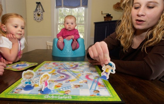 Michelle Hastings, right, plays Chutes and Ladders with her daughter Peyton Hastings, 2, left, as her daughter Andersen, 5 months, center, watches at their home in Holliston, Mass.