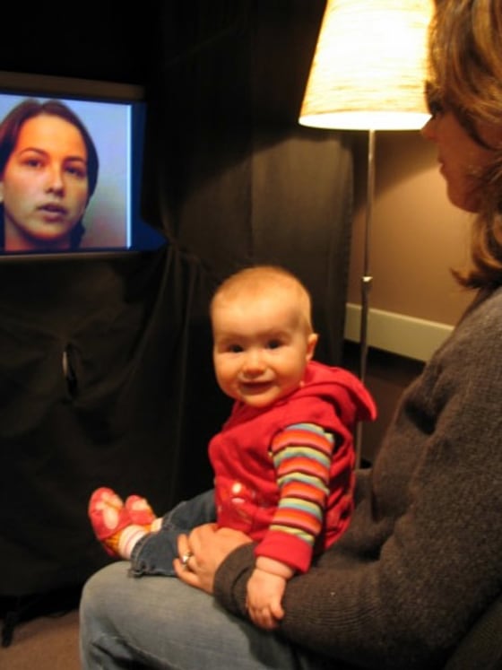 A baby sits on a caregiver's lap before a television screen used in a University of British Columbia study to see how babies watch the silent faces of bilingual speakers reciting sentences in both English and French. The caregiver wears darkened sunglasses to prevent influencing the baby.