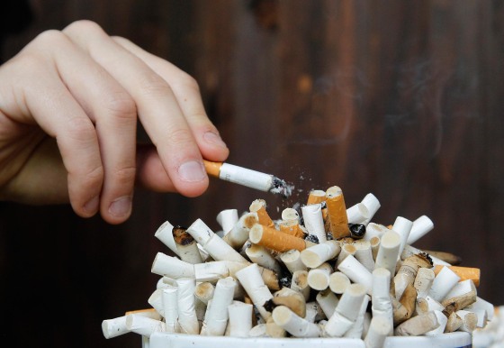 Image: A man taps ashes off his cigarette into an ashtray filled with cigarette butts on a table.