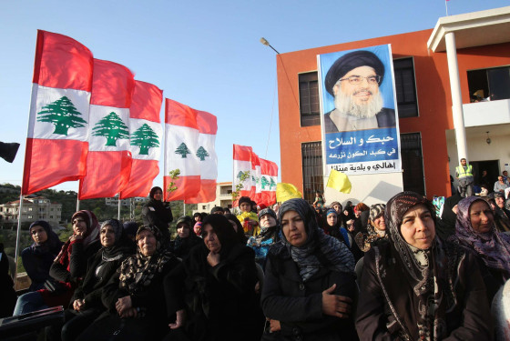 Image: Hezbollah supporters stand in front of a banner of their leader Nasrallah as they listen to his speech in Aineta