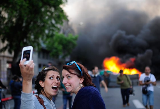 Tourists take a selfie as demonstrators burn a trash container during a May Day rally in Barcelona, Spain, on May 1, 2014.