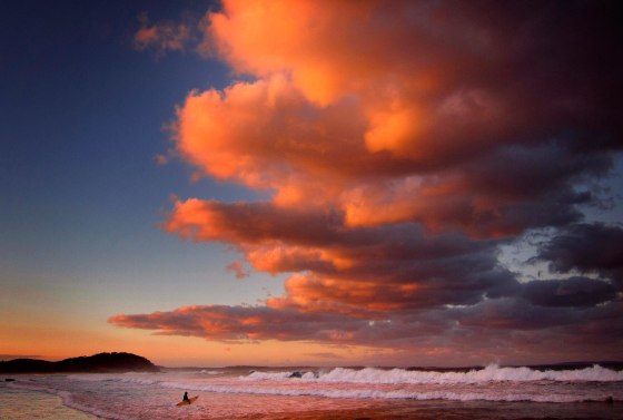 Image: Surfer holding her board wades through the surf as clouds above are lit by the setting sun at Mollymook Beach on the south coast of New South Wales