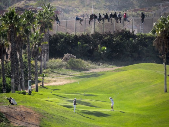 Image: Golfers playing from a fairway back-dropped by African migrants