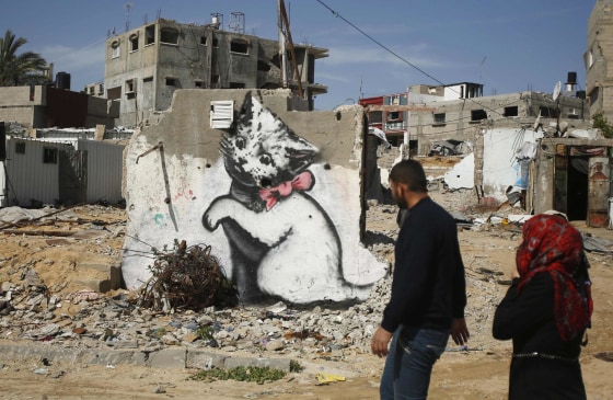 Image: Palestinians walk past a mural of a kitten, presumably painted by British street artist Banksy, on the remains of a house in Biet Hanoun