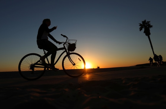 Image: A woman looks at her phone as she rides her bike along the boardwalk in Venice Beach