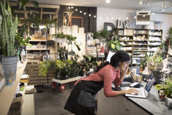 Image: Female shop owner talking on cell phone working at laptop at plant shop counter