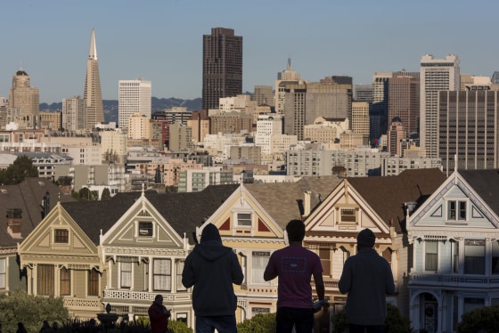Image: The silhouettes of pedestrians stand in front of Victorian homes and the downtown skyline in San Francisco