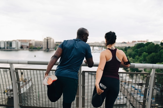 Image: Rear view of male and female athletes stretching legs while standing on footbridge over sea in city