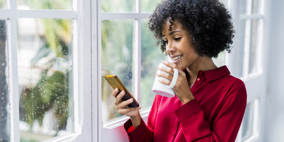 Smiling woman with cup of coffee and cell phone standing at the window at home