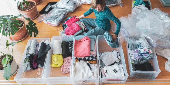 Woman sitting on her floor surrounded by clothes, organizing drawers and big storage bags for under her bed