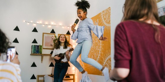Girl in her bedroom, jumping on her bed, holding a brush pretending to sing, surrounded by three friends