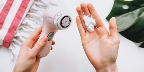 Image of a Woman holding a pink facial cleansing brush in one hand and soap in the other hand