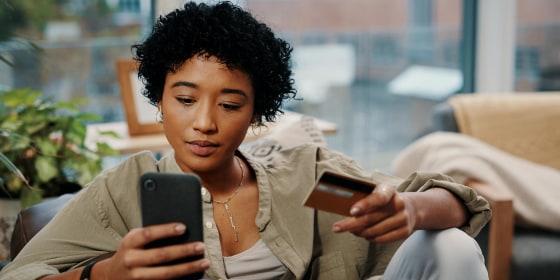 Shot of a young woman holding her credit card while using her cellphone at home