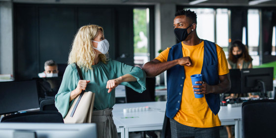 Two coworkers inside an office wearing face masks, bumping elbows
