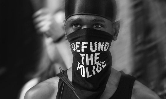 Image: A Black trans liberation activist wears a bandana that reads \"Defund the Police\" in New York City.