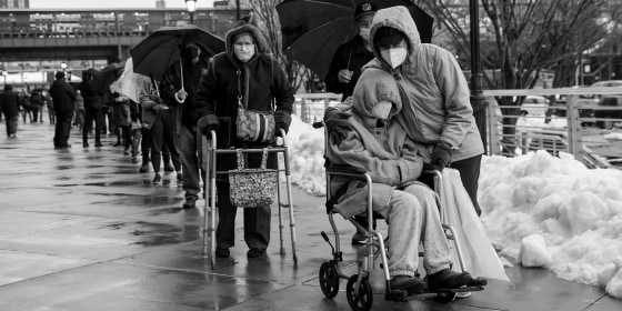 Image: A person in a wheelchair in a line of people lined up outside a Covid-19 vaccination site.
