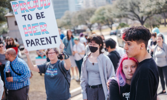 Image: A demonstrator holding a sign that reads,\"Proud to be be her parent\". The words, \"Proud\" and \"her\" are in the transgender pride flag colors.