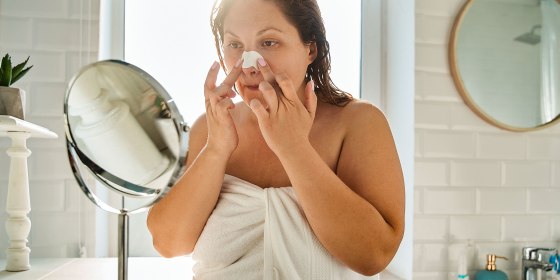 Woman applying nose strips while looking at mirror. Concept of face skin care. Young european girl wearing towel. Interior of bathroom in modern apartment