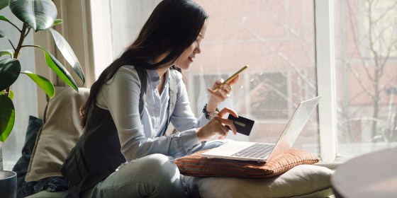 A middle-aged asian woman in blue jeans sitting on the bed in a yoga pose in front of a laptop. Remote work at home. Online shopping in quarantine period
