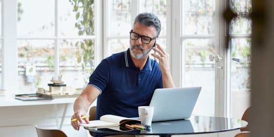Mature businessman using mobile phone while writing notes at table in office