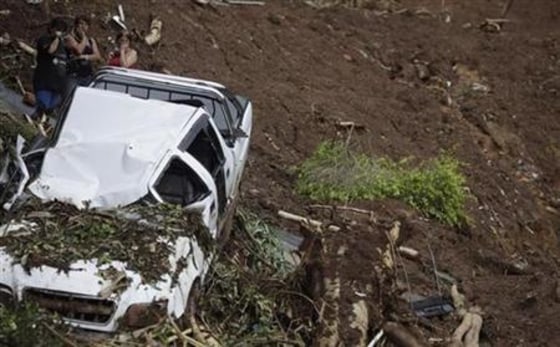 People watch behind a wrecked car as rescue workers look for bodies after a landslide in Nova Friburgo
