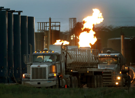 Drivers and their tanker trucks capable of hauling water and hydraulic fracturing liquid line up near a natural gas burn off flame and storage tanks in Williston, N.D., on June 9, 2014.