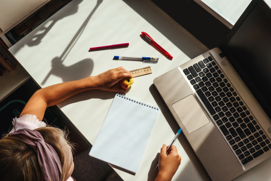 A girl seated at a desk with a laptop, notebook and pens.