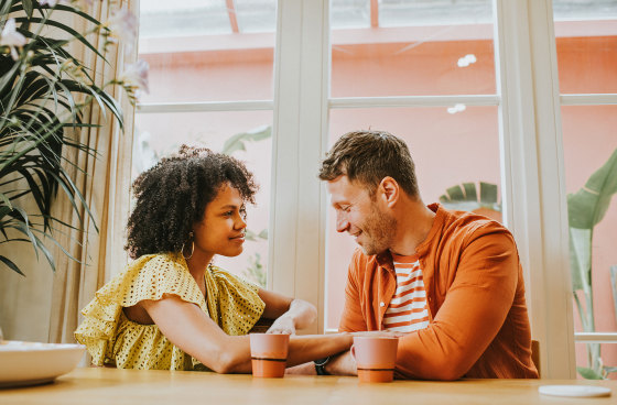 A young couple sit at a dining table sipping hot drinks and flirt.