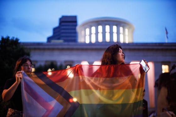 Two transgender rights advocates hold a large progress pride flag in front of the Ohio Statehouse
