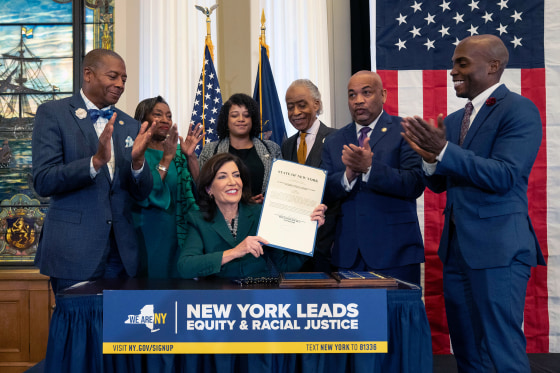 James Sanders, NY State Senator; Andrea Stewart-Cousins, Majority Leader of the NY State Senate; Michaelle Solages, NY State Assembly Woman; Rev. Al Sharpton; Carl Heastie, Speaker of the NY State Assembly; Dr. Yohuru Williams, Founding Director of the Racial Justice Initiative at the University of St. Thomas applaud as Kathy Hochul holds the signed bill.