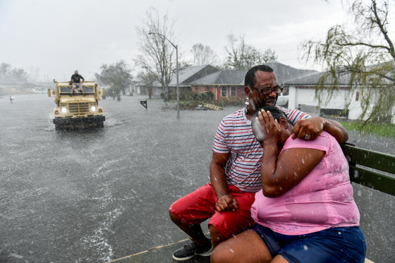 Two people embrace while sitting on a bench as a rain shower soaks them outside, as a volunteer evacuation truck is in the distance