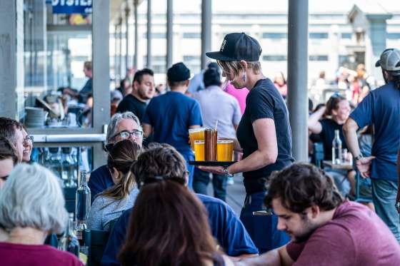 Customers are seated at a restaurant outdoors, a server walks through carrying drinks