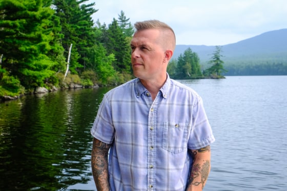 Jared Golden stands outside in front of a body of water lined with trees